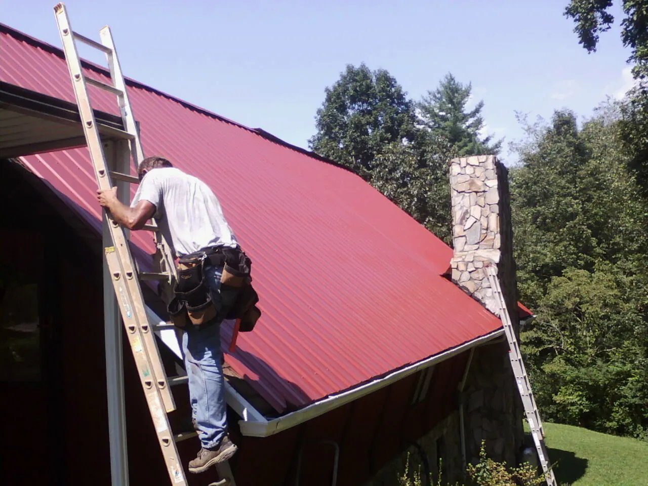 Roofer on a ladder beside a red metal roof and stone chimney — Soffit & Fascia Repair by Roofing Brothers in Great Barrington